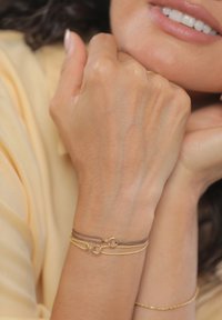 Zapestnice v zlati barvi na zapestju, ena tanka zlata verižica in en rjavi kabel, obe s krožnimi poudarki. Gladka koža in svetla tkanina v ozadju.