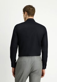 Man draagt een zwart overhemd met lange mouwen en grijze broek, staat met zijn rug naar de camera tegen een effen witte achtergrond.