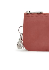 Funda con cremallera en suave tela de color óxido. Presenta herrajes en plata y un charm metálico de mono sujeto por un llavero. Textura suave.
