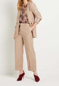 Costume beige ajusté avec un pantalon large et un blazer, superposé sur un haut à motif floral. Des chaussures à talons hauts bourgogne complètent le look.