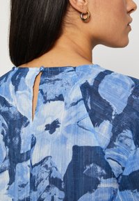 Top blu con motivo a fantasia e schiena con apertura a goccia, realizzato in un tessuto testurizzato. Il design presenta tonalità chiare e scure di blu con motivi astratti.