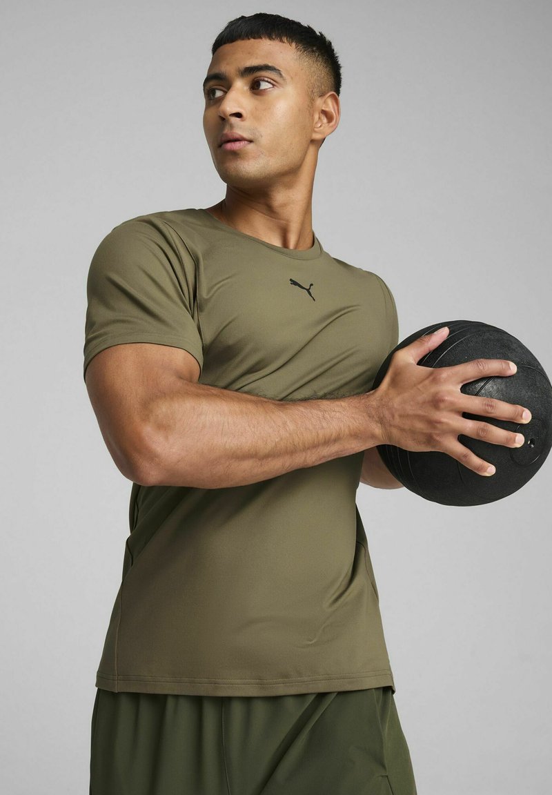 Camiseta atlética verde oliva de manga corta, con un logo negro en el pecho; sosteniendo una pelota medicinal negra texturizada.
