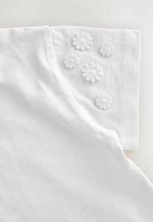 T-shirt en coton blanc avec des manches repliées ornées de six motifs floraux brodés. Texture lisse, design simple avec des lignes épurées.