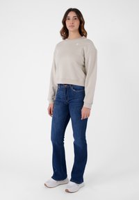 Maglione cropped beige con una texture a costine, jeans a zampa d'elefante blu e sneakers bianche con suola texturizzata, ambientati su uno sfondo bianco semplice.