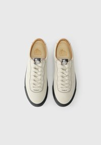 Bēša auduma kedas ar melnu gumijas zoli, apaļu purngalu un sešām acīm. Melnu diegu detaļas un logo uz mēles un iekšējās zoles.