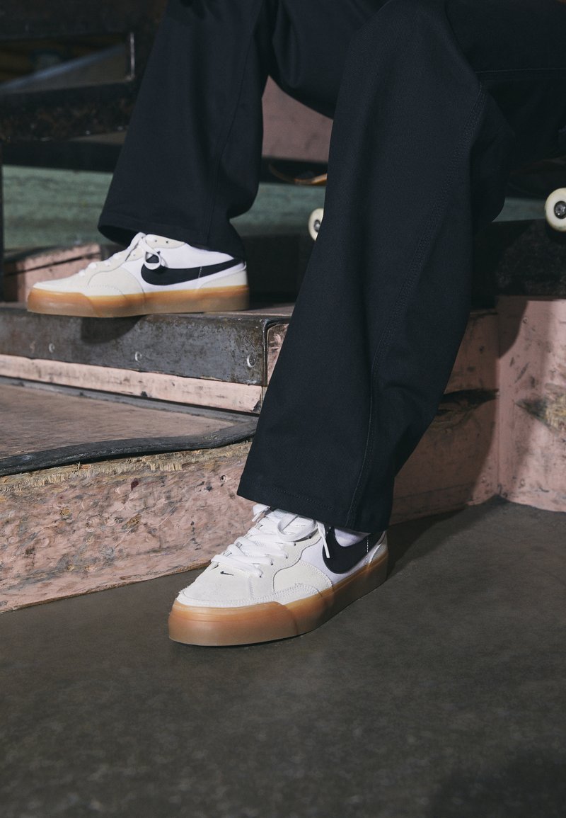 Zapatillas blancas con acentos en negro, con suela de goma, parte superior de ante y cuero texturizado, y cordones planos, colocadas en un escalón.