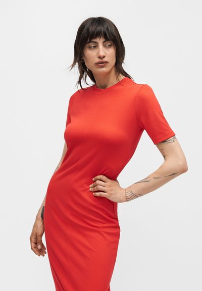 Γυναίκα με κοντά σκουρόχρωμα μαλλιά, φορώντας εφαρμοστό φωτεινό κόκκινο φόρεμα με κοντά μανίκια, ποζάρει με το ένα χέρι στη μέση της, με ορατά τατουάζ στα χέρια.