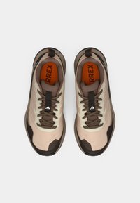 Μπεζ αθλητικά παπούτσια με σκούρες καφέ λεπτομέρειες, υφασμένο πάνω μέρος, παχιά λάστιχη σόλα και γκρι κορδόνια· διαθέτουν πορτοκαλί εσωτερική σόλα και ανακλαστική λεπτομέρεια.