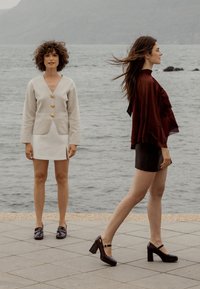 Deux modèles se tiennent près de l'eau. L'un porte un cardigan léger avec des boutons dorés et une jupe blanche ; l'autre, un haut à volants bordeaux et un short noir.