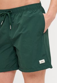 Mørkegrønne svømmeshorts lavet af letvægtsstof, med elastisk talje med snor, sidelommer og et lille logo mærke.