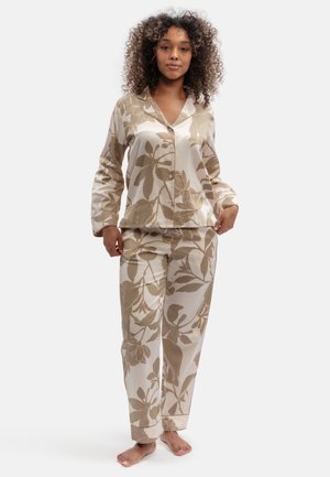 Femme aux cheveux bouclés portant un haut beige et crème à motifs floraux, boutonné, à manches longues et un pantalon assorti, debout pieds nus.