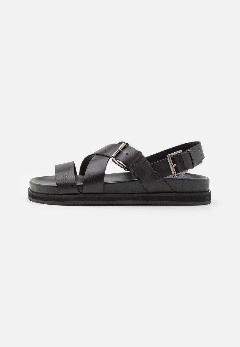 Schwarze Ledersandalen mit Kreuzriemen-Design, verstellbarer Schnalle, flacher Sohle und glatter Oberfläche. Seitenansicht.