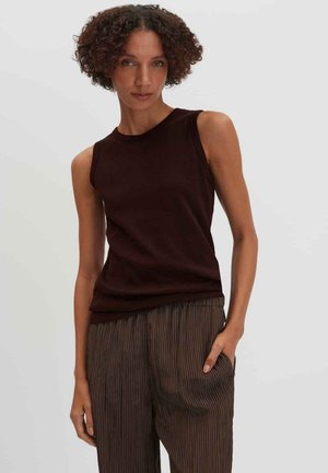 Vrouw met krullend haar, gekleed in een mouwloze donkerbruine top en loszittende bruine gestreepte broek tegen een effen lichte achtergrond.