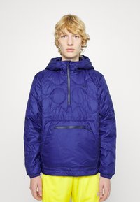 Giacca puffer blu con cappuccio, dotata di un'apertura frontale con mezza zip e una tasca con zip. I pannelli testurizzati creano un motivo trapuntato.