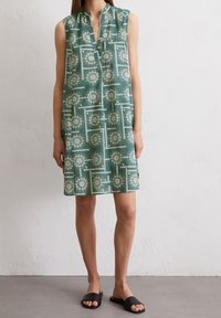 Mintgrön ärmlös klänning med placket-halsringning, vita cirkulära mönster och lös passform. Matchas med svarta slip-in sandaler.