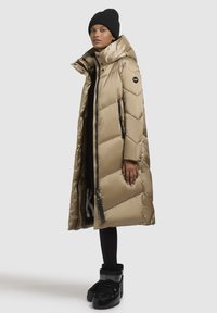 Long puffer coat in metallic beige with chevron quilted design, high collar, and black zipper pockets. Paired with black leggings and boots.