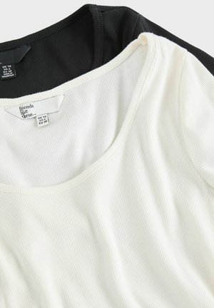 Zwei gerippte Tanktops in Schwarz und Weiß, mit weitem Halsausschnitt und weichem Stoff. Etiketten sichtbar mit Größeninformationen.