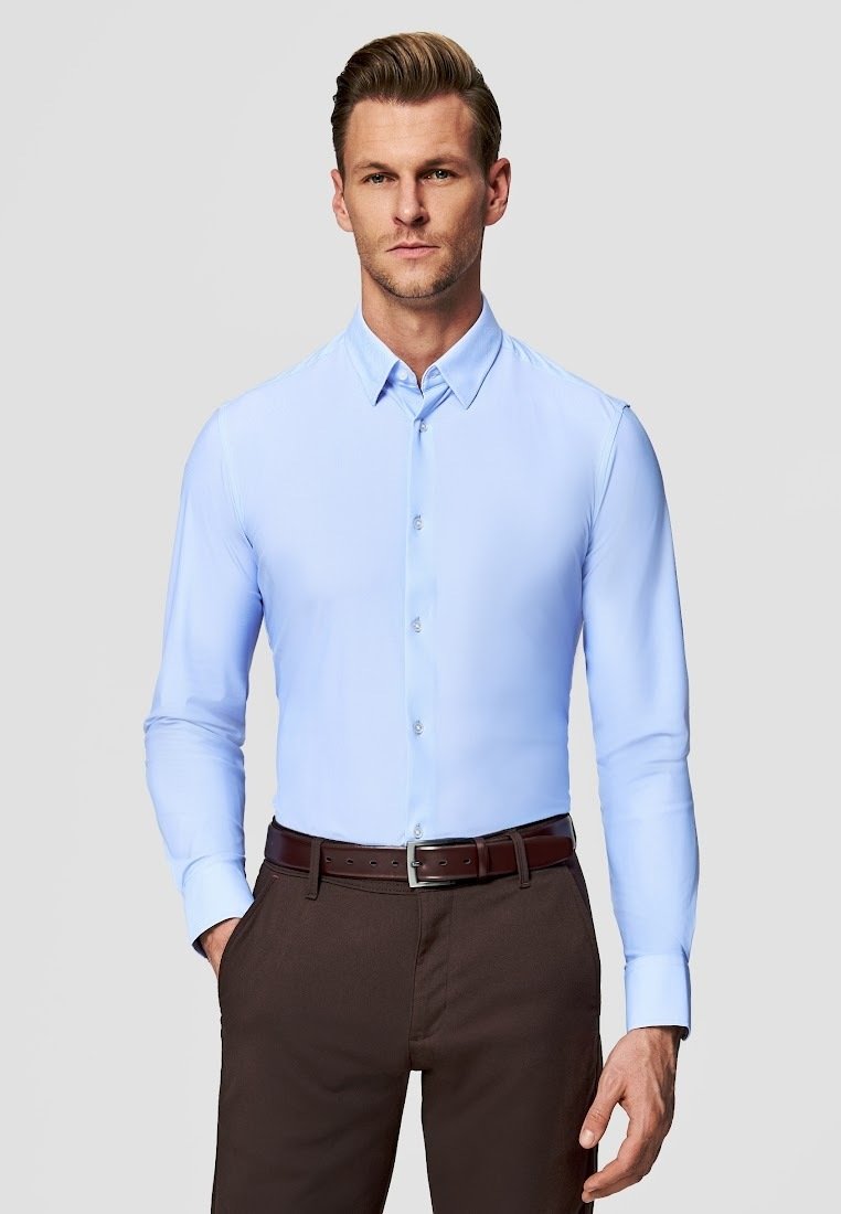 Mężczyzna o krótkich włosach, ubrany w jasnoniebieską koszulę zapinaną na guziki, wpuszczoną do ciemnobrązowych spodni z ciemnobrązowym paskiem, stojący z jedną ręką w kieszeni.