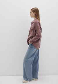 Ein Model trägt ein staubiges Rosafarbenes Cordhemd mit Tasche und hellblauen, weit geschnittenen Jeans. Der Stoff wirkt weich und hat eine lässige Passform.