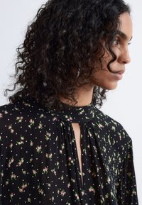 Schwarze Bluse mit kleinen rosa Blumenmustern und weißen Punkten. Sie hat einen hohen Kragen und ein Schlitzdetail am Kragen. Leichter Stoff.