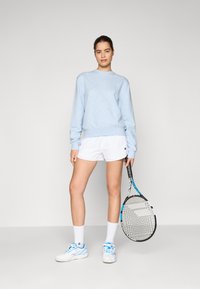 Light blue sweatshirt, white shorts, white socks, and blue-accented tennis shoes. Holding a tennis racket with a black and white frame.