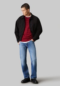 Giacca nera con zip sopra un maglione bordeaux, stratificata con una maglietta bianca, abbinata a jeans azzurri chiari e scarpe nere.