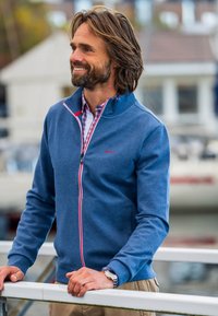 Blauwe sweatshirt met rits en rode en witte accenten; zachte stof, hoge kraag en zijzakken. Draagt over een geblokt shirt.