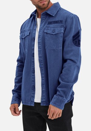 Chemise en denim bleu avec un devant à boutons, deux poches poitrine et un patch sur la manche gauche. Texture lisse, col standard et coupe décontractée.