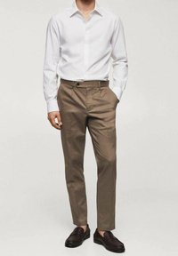 Chemise blanche à manches longues avec boutons, associée à un pantalon beige. Le pantalon a une texture lisse et un détail de bouton unique à la taille.