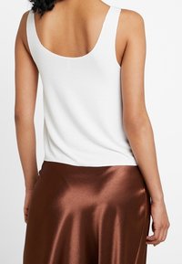 Bílý žebrovaný top bez rukávů s širokými ramínky, který má výstřih vzadu; v kombinaci se hnědou saténovou sukní s hladkým povrchem a mírným leskem.