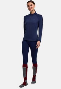 Marineblauw top met lange mouwen en een halve ritssluiting, met een logo op de mouw, gecombineerd met nauwsluitende marineblauwe legging en kniekousen in rood en grijs met een patroon.
