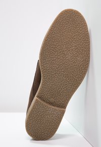 Semelle de chaussure en cuir suédé marron avec un dessous en caoutchouc texturé. Forme arrondie, présentant un motif tactile pour une meilleure adhérence. Le côté montre un détail de couture minimal.