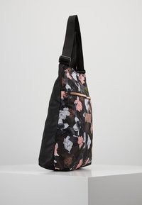 Mustasta olkalaukusta, jossa on vaaleanpunaisia, valkoisia ja ruskeita kukkakuoseja, etupuolella on vetoketjullinen tasku ja säädettävä leveä musta olkahihna, joka on asetettu valkoiselle pedestaalille.