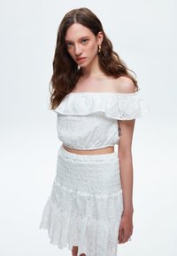 Weißes Off-Shoulder-Oberteil mit gerüschtem Ausschnitt und floraler Stickerei, kombiniert mit einem passenden Smocking-Rock mit gestuften Rüschen.