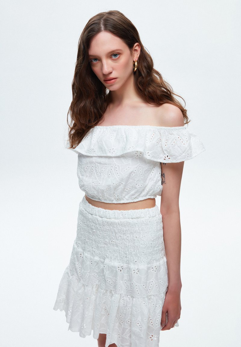 Weißes Off-Shoulder-Oberteil mit gerüschtem Ausschnitt und floraler Stickerei, kombiniert mit einem passenden Smocking-Rock mit gestuften Rüschen.