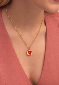 Goldene Kettenanstecknadel mit einem runden Anhänger, der ein rotes Herz und eingravierte Akzente zeigt, getragen auf einem rosa Stoffoberteil.