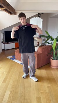 Czarna koszulka polo z kołnierzykiem i detalem na guziki trzymana przez osobę w pomieszczeniu z drewnianą podłogą i pomarańczowo-paskową kanapą.