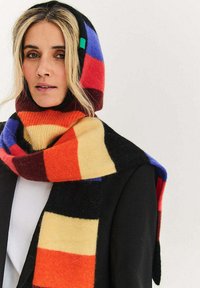 Wielokolorowy wełniany szalik z kapturem, z dużymi geometrycznymi pasami w kolorach czerwonym, pomarańczowym, czarnym, niebieskim i żółtym. Miękka tekstura, wyraźna konstrukcja.