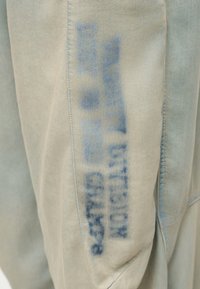 Αχνό μπλε στένσιλ κείμενο που γράφει «ΕΡΕΥΝΑ ΤΗΣ ΑΣΤΥΝΟΜΙΑΣ ΓΙΑ ΕΓΚΛΗΜΑ» σε ραφή ανοιχτού μπεζ υφάσματος.