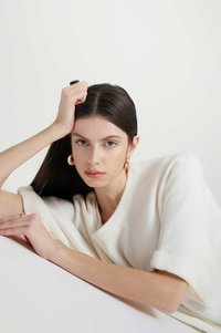 Maglione bianco lavorato a maglia con maniche arrotolate, texture morbida, modella che posa con capelli scuri e orecchini a cerchio dorati, sfondo neutro.