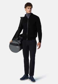 Sudadera negra con cremallera y un diseño ajustado, combinada con pantalones azul marino y zapatillas azul marino. Sosteniendo un bolso de duffle gris circular etiquetado "45L".