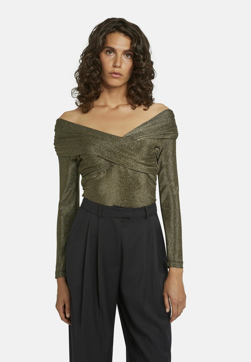 Langärmlige, schulterfreie Bluse aus metallisch olivgrünem Stoff mit strukturierter Drapierung. Kombiniert mit hochgeschnittenen schwarzen Hose.