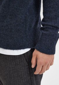 Marineblauer Wollpullover mit geripptem Saum und Bündchen, mit einer strukturierten Oberfläche. Getragen über einem weißen Hemd, kombiniert mit dunkelgrauen Hosen.