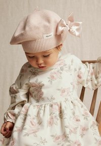 Ljusrosa stickad beret med en satinrosett, bärs av ett barn i en klänning med blommönster som har rosa rosor och gröna blad.