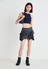 Top cropped em malha azul-marinho com detalhes em prateado, combinado com uma saia mini cinza em camadas. A modelo usa botins pretos e segura uma bolsa azul com padrão.