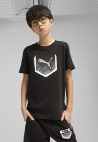 Camiseta de algodón negra con un gran logo de Puma en blanco dentro de un diseño de escudo punteado. Combinada con pantalones negros a juego que muestran el logo.