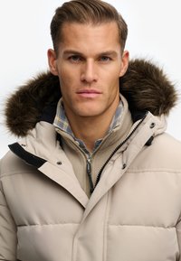 Beżowa kurtka puchowa z czarnym kapturem, ozdobiona sztucznym futrem. Nałożona na brązową polarową bluzę z zamkiem i niebieską koszulę w kratę.