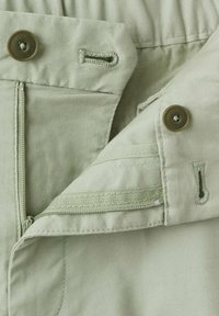 Primer plano de pantalones verde claro que muestran botón, ojal y cremallera parcialmente abierta.