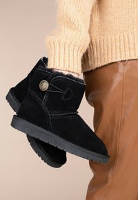 Botas de ante negras hasta el tobillo con puntera redonda, forro de oveja, botón decorativo y suela de goma texturizada, sostenidas frente a un fondo beige.