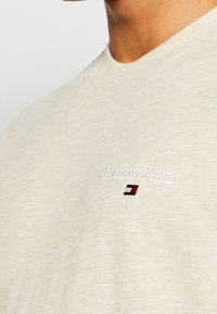 Béžové bavlněné tričko s kulatým výstřihem, které zdobí bílé vyšívání "Tommy Jeans" a malá nášivka v červené, bílé a modré barvě.